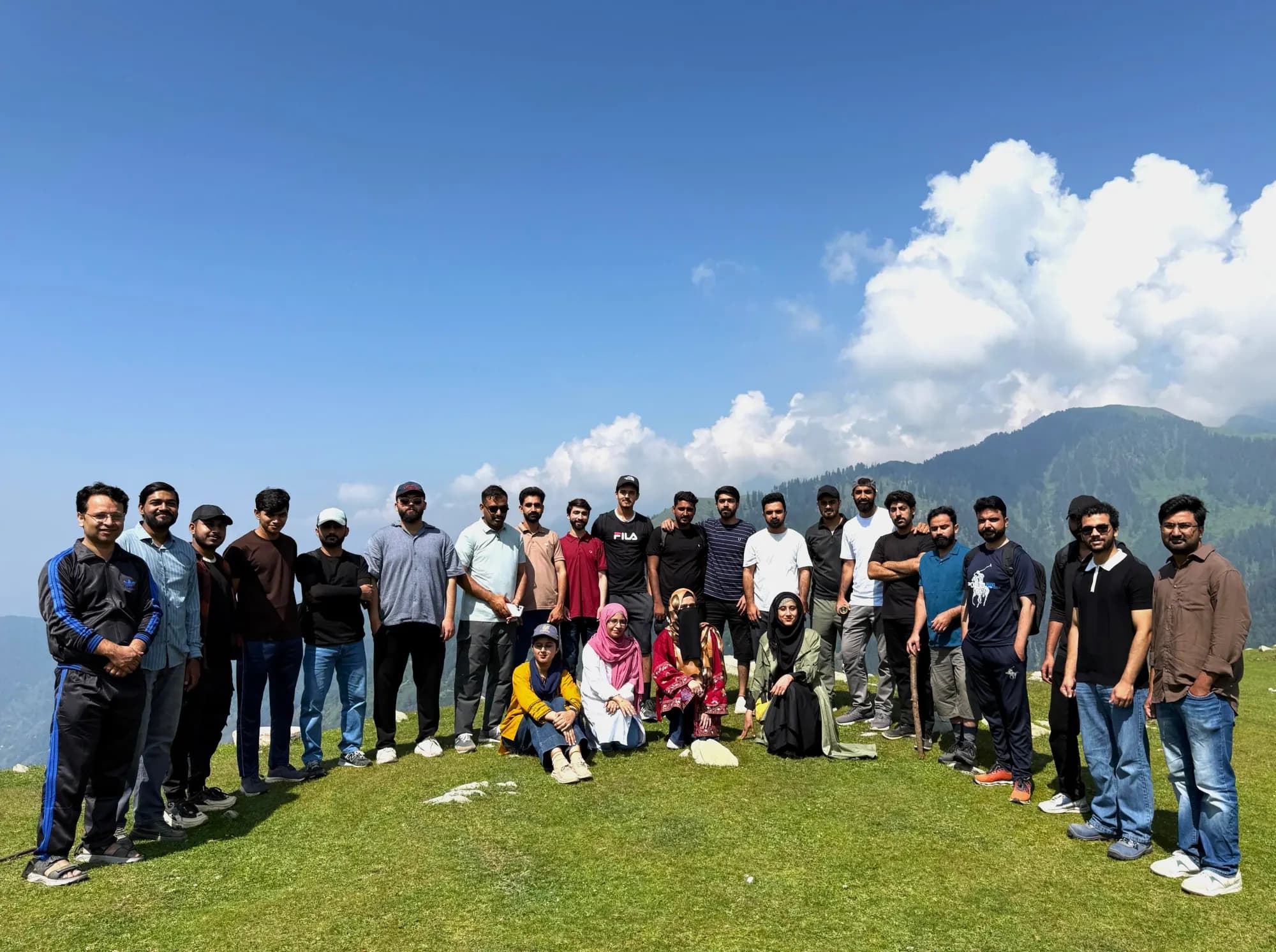 The team standing together on a grassy mountaintop under a blue sky, with forested peaks and clouds behind